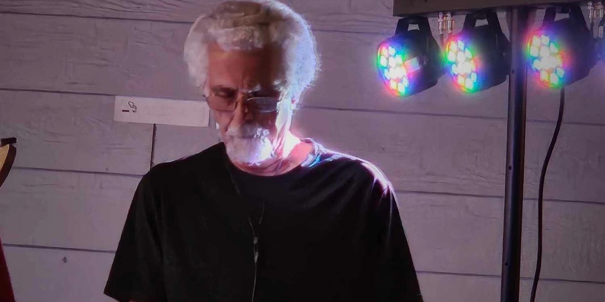 Elderly man with white hair and glasses illuminated by colorful lights.