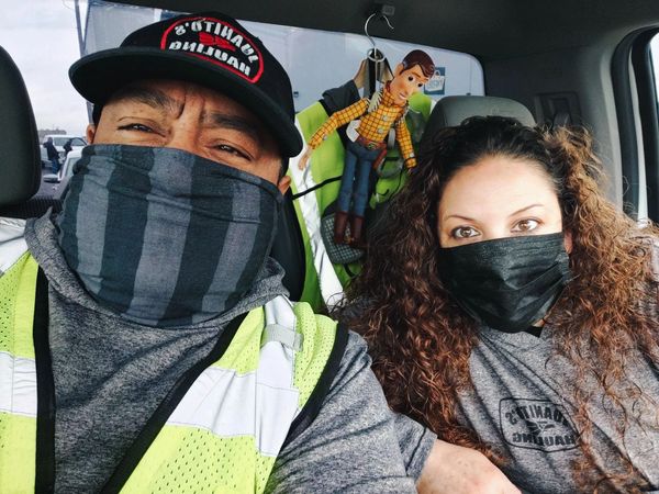 Two masked people sitting in a car with a Woody doll in the background.