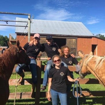 Four people in black shirts posing with two horses near a barn on a sunny day.