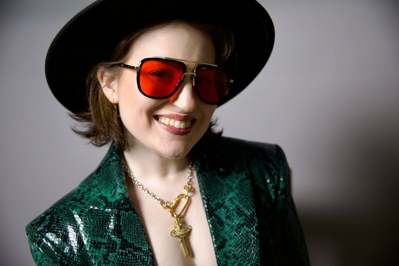 Closeup of Hillary with green jacket, red lens glasses, hat, and key necklace, smiling