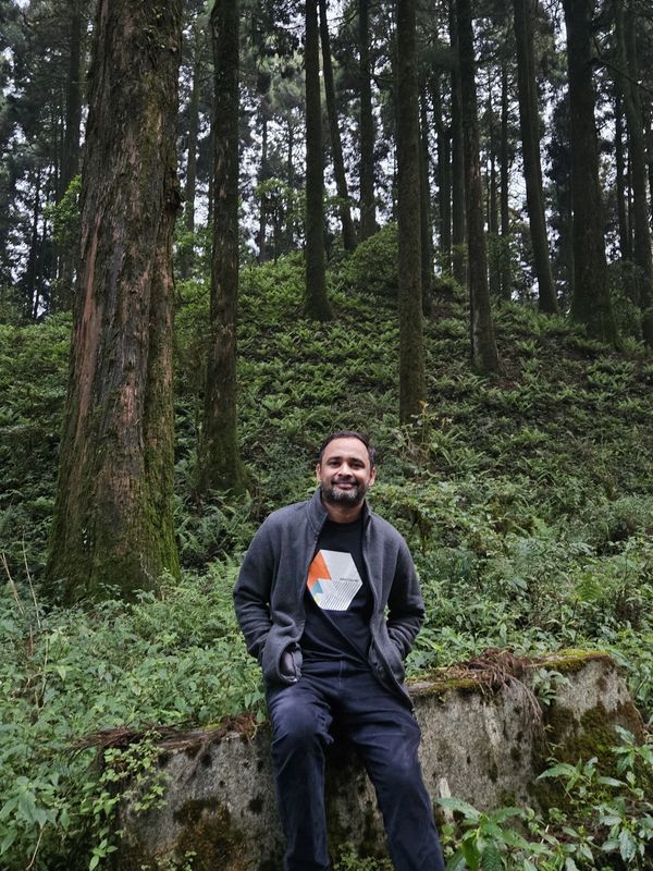 Man sitting on a mossy stone in a dense forest, smiling.