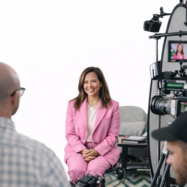 A woman in a pink suit smiles during a filmed interview with crew members.
