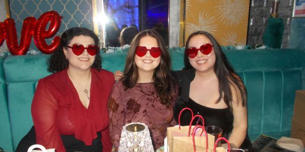 Three women wearing red heart-shaped sunglasses smiling at a party.