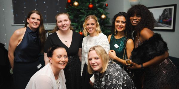 A group of seven women dressed elegantly, posing happily in front of a decorated Christmas tree.