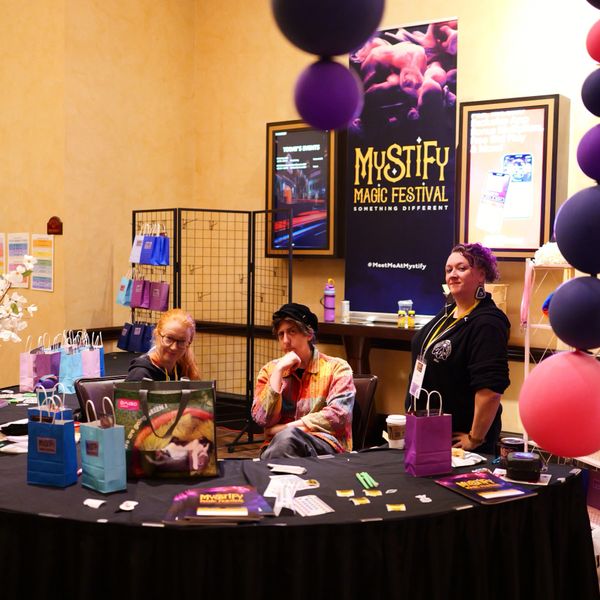 Three women at a Mystify Magic Festival booth with colorful bags and balloons.