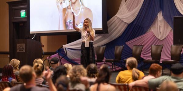 Woman speaking to an attentive audience at a conference with a large screen behind her.
