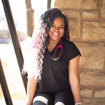 Smiling woman with long curly hair and a stethoscope, sitting against a stone wall.