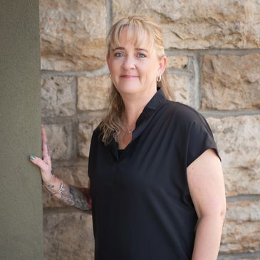 Smiling woman in black shirt standing by a stone wall.