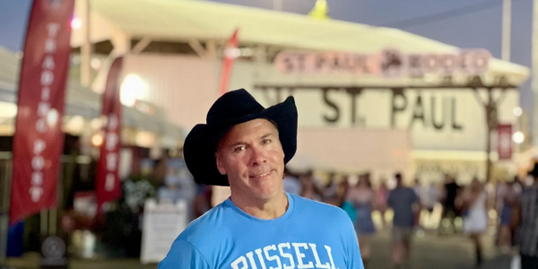 Man in a cowboy hat and blue Russell Athletic shirt at an outdoor event.