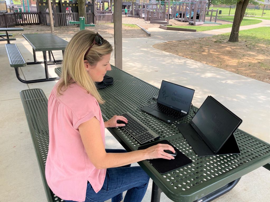 Woman working outdoors on laptops at a park picnic table.