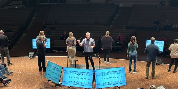 A conductor leads a choir rehearsal with singers spaced on stage and music displayed on screens.