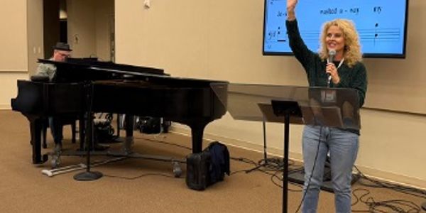 Woman speaking with microphone beside pianist in a music room.
