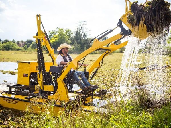 Man operating a yellow excavator removing plants from water.