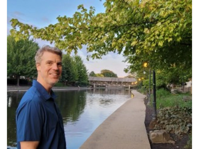 Man smiling by a peaceful riverside path with greenery and a covered bridge.