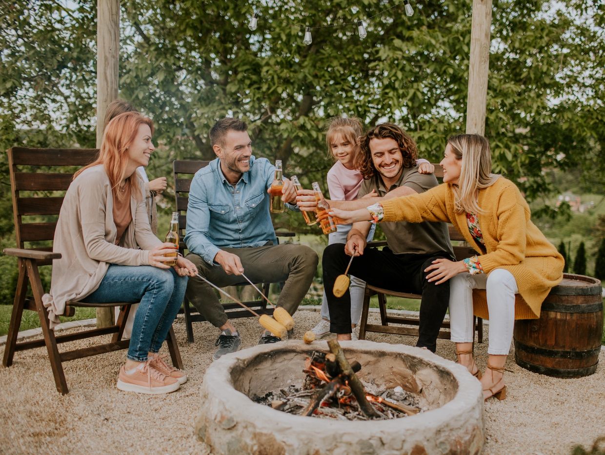 Friends enjoying a bonfire, roasting marshmallows, and toasting with drinks outdoors.