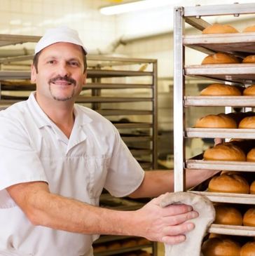 Baker smiling while handling fresh bread on racks in bakery.