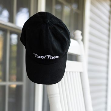 A black hat from our pronoun collection which features white text that reads they/them