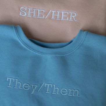 Three of our pronoun crewnecks in blue, pink, and grey featuring he/him, she/her, and they/them