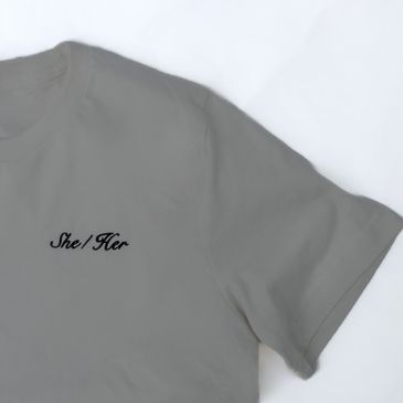 A grey t-shirt from our pronoun collection which features cursive font reading she/her in black