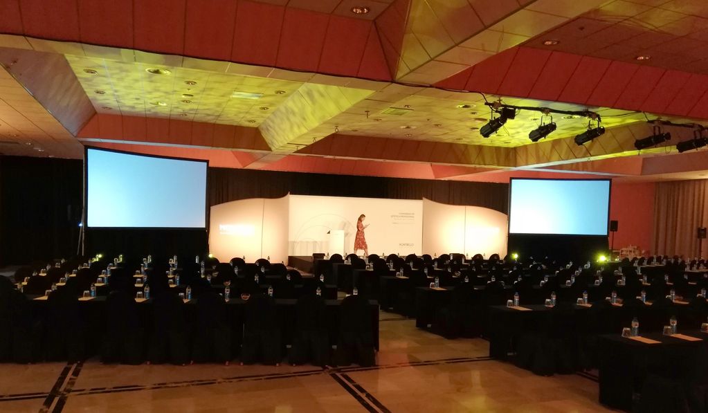 Conference room with rows of chairs and two large screens, a person on stage.