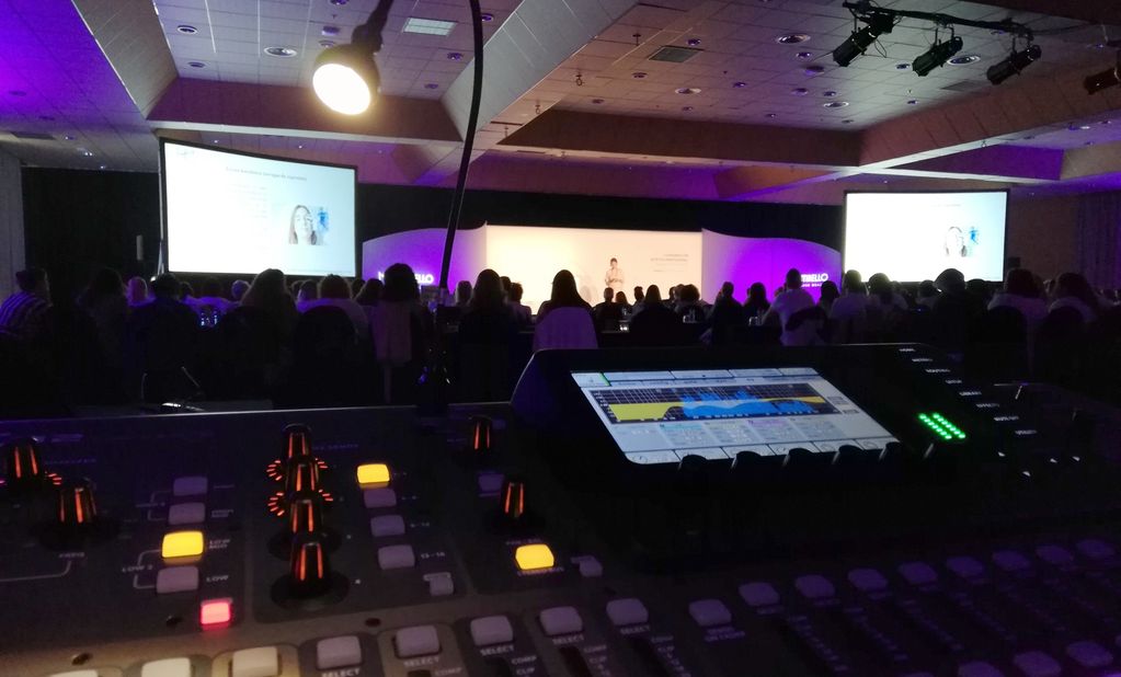 Audio mixing console in the foreground at a conference with audience and presentation screens.