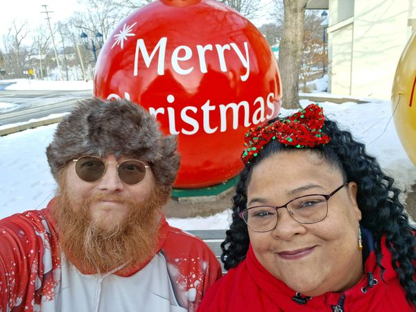 Two people in festive attire smiling in front of a large red Christmas ornament.