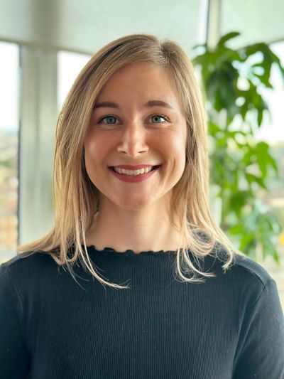 Smiling woman with blonde hair in front of a window and green plant.