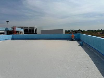 Worker painting blue waterproof coating on rooftop edges under cloudy sky.