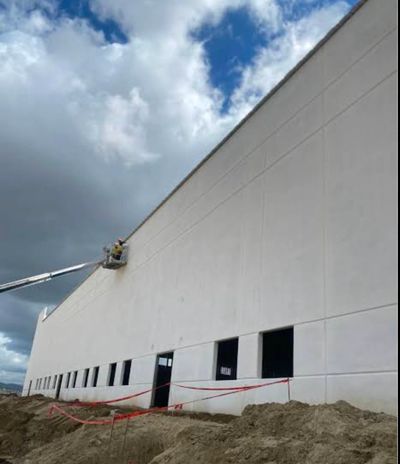 Workers on a lift painting or inspecting a large industrial building under cloudy skies.