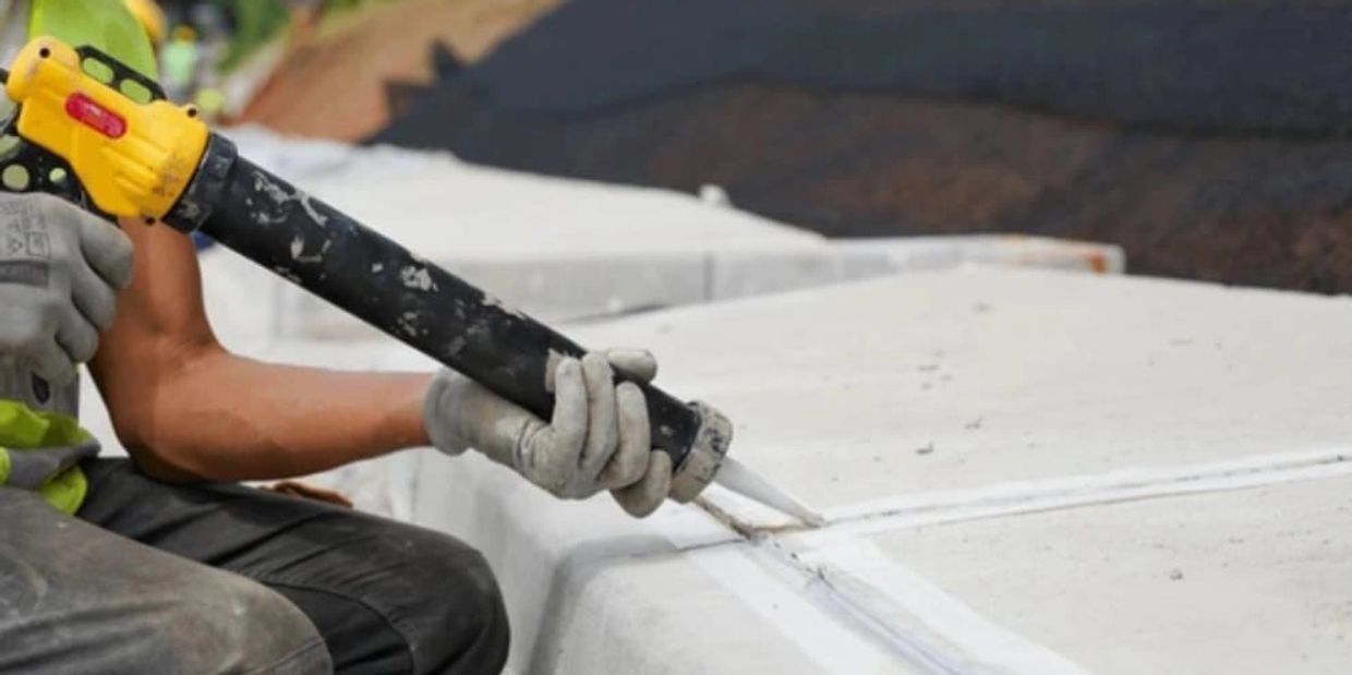 Worker applying sealant to concrete joints with a caulking gun outdoors.