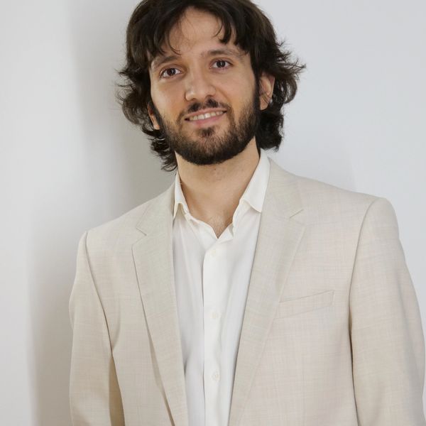 Smiling man with medium-length hair and beard, wearing a light beige suit jacket.