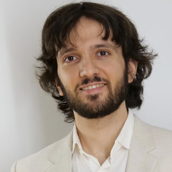 Smiling man with medium-length hair and beard, wearing a light beige suit jacket.