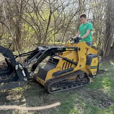 Person operating a yellow Vermeer mini skid steer loader in a wooded area.