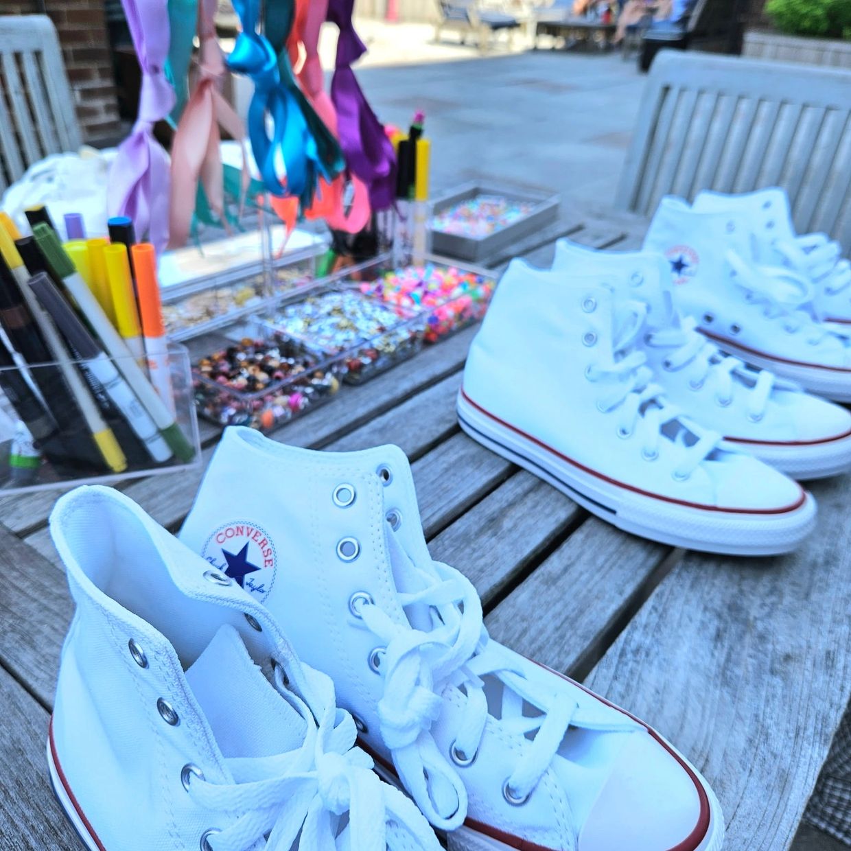White Converse shoes lined up on a wooden table with colorful craft supplies in the background.