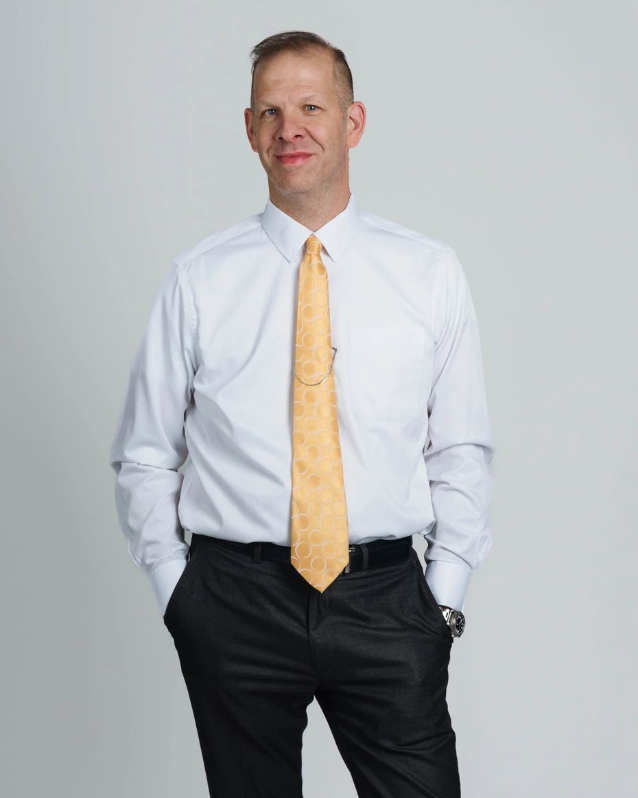 A man in formal attire stands confidently with hands in pockets against a plain background.