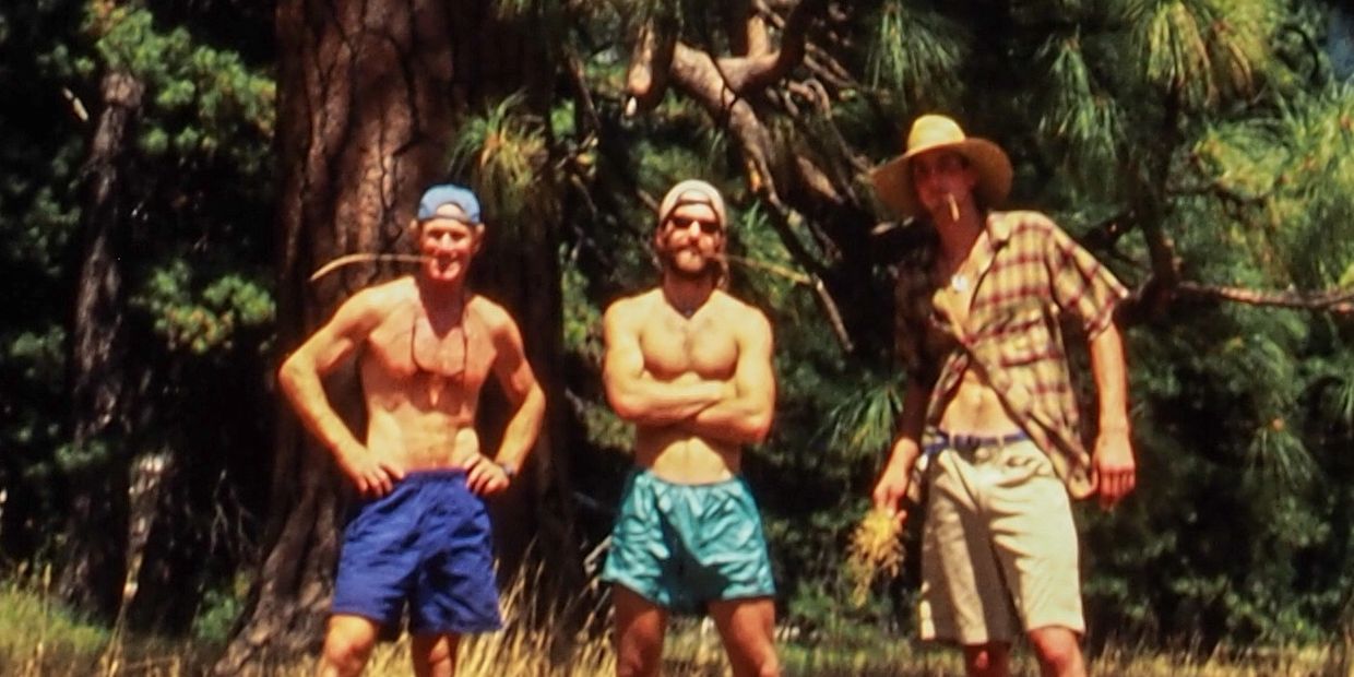 Three shirtless men in shorts stand confidently in a forest clearing.
