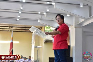 A woman in a red shirt speaking at a podium indoors.