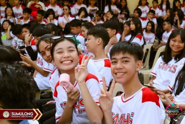 Smiling students in red and white shirts at a school event, making peace signs.