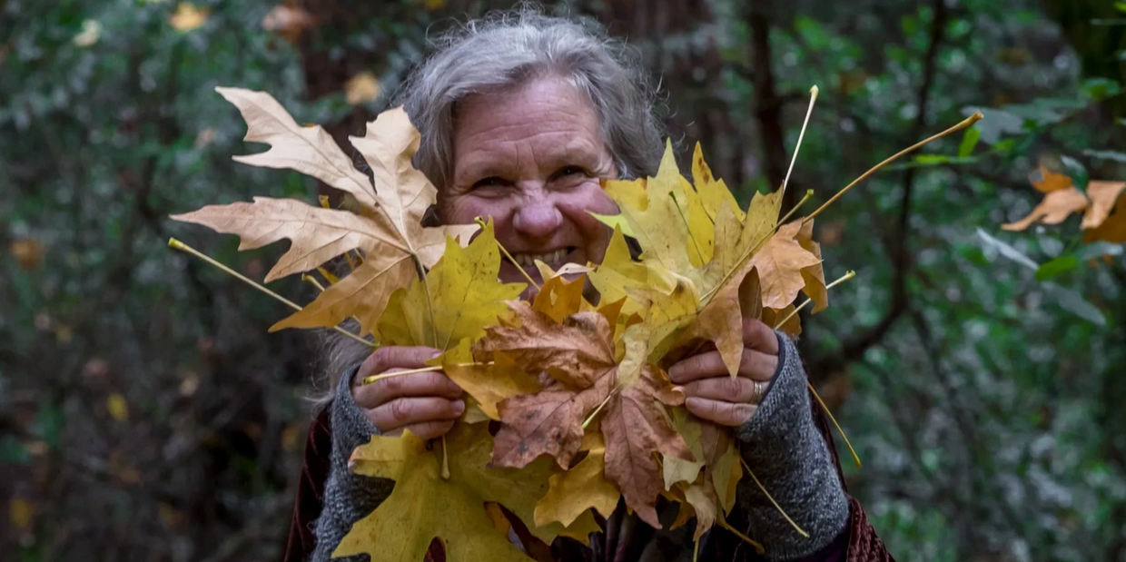 Elderly woman joyfully holding autumn leaves in a forest.