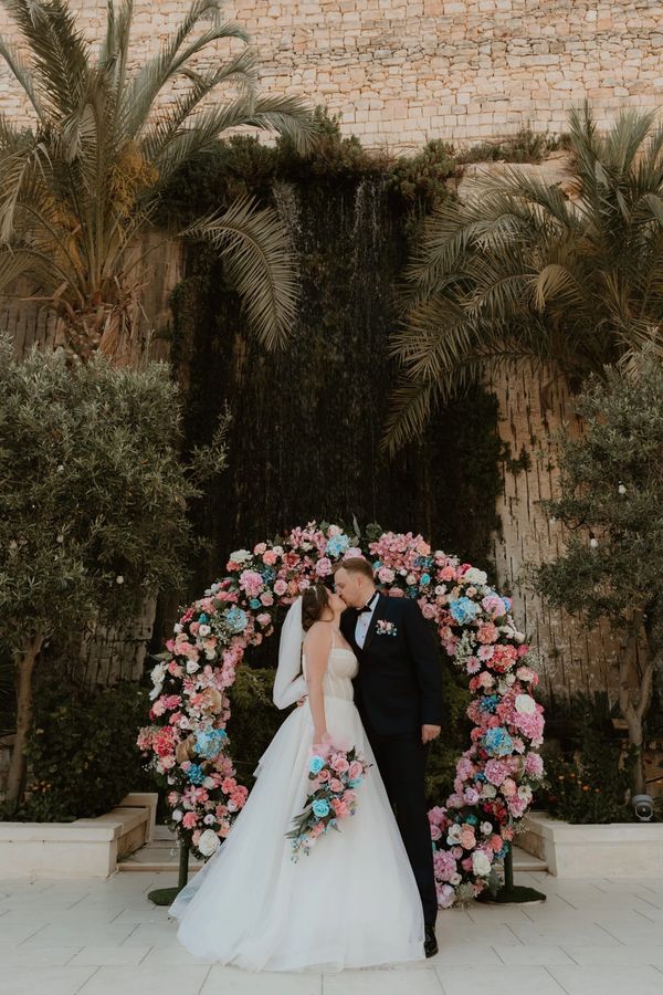Bride and groom kiss in front of a floral arch at their wedding.