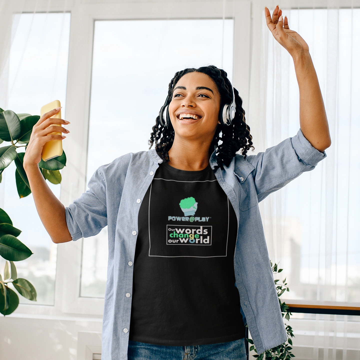 Joyful woman dancing with headphones, holding a phone, wearing a motivational Power@Play t-shirt.