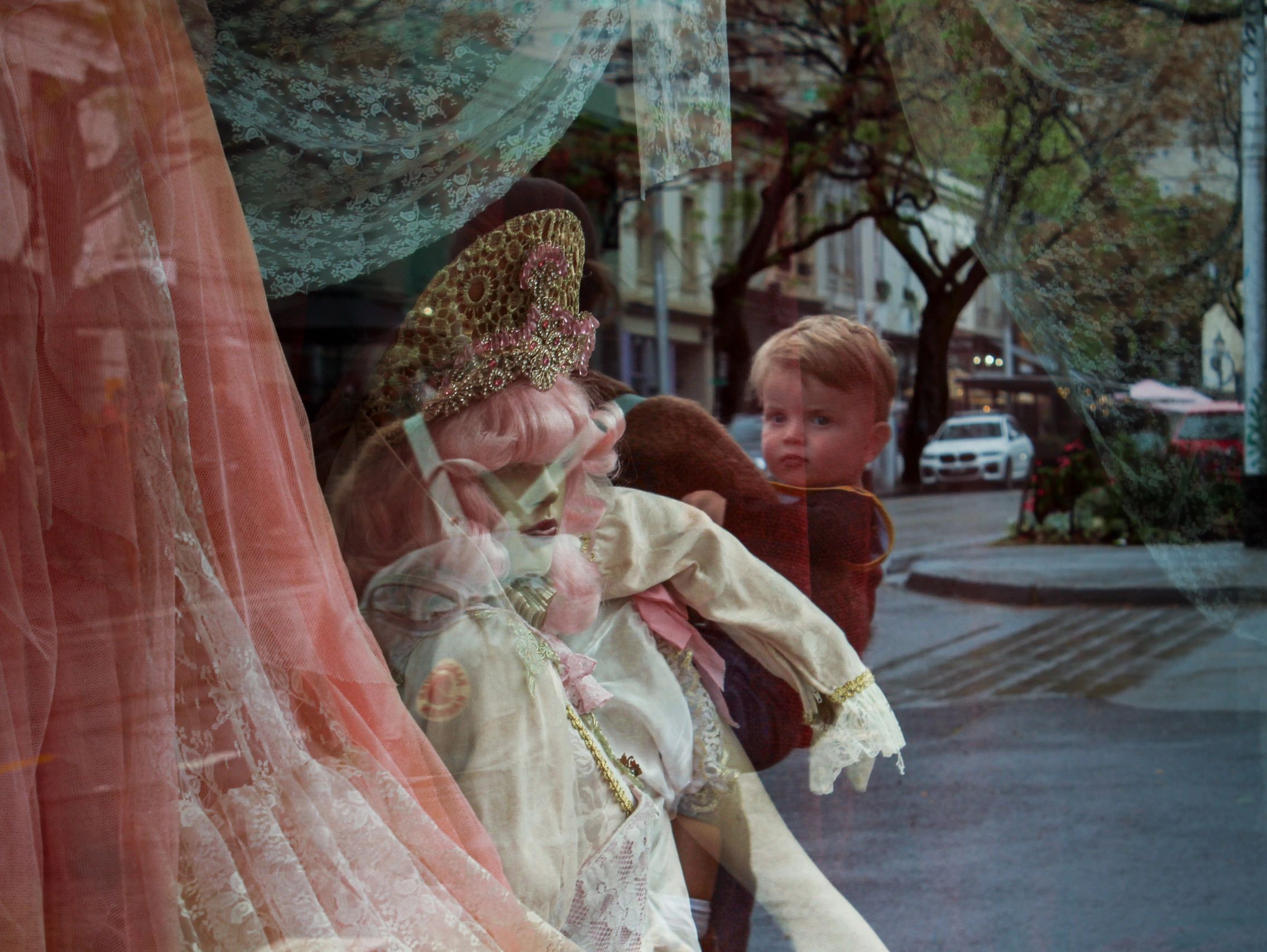 Reflections of a child and a doll in a vintage outfit through a shop window.