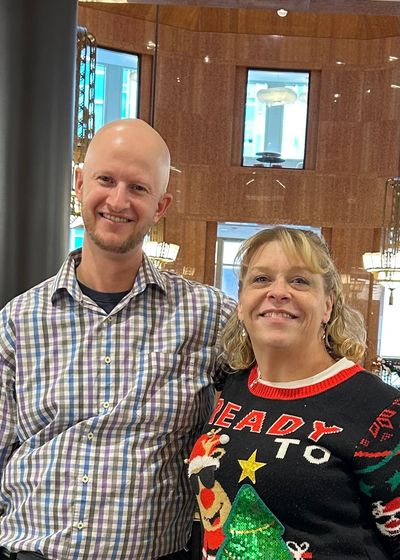 Two people smiling indoors, one wearing a festive Christmas sweater.