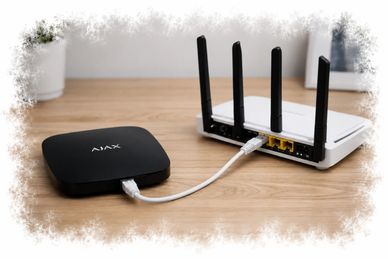 A black AJAX device connected to a white router on a wooden table.