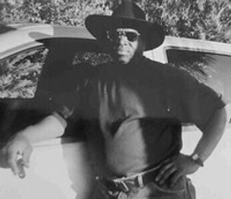 Man in black hat and sunglasses leaning on car.