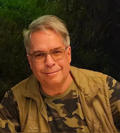 Smiling man wearing glasses and camouflage clothing outdoors at night.