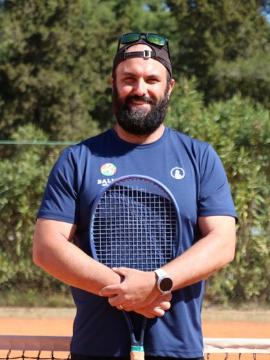 Bearded man in a cap holding a tennis racket on a sunny tennis court.