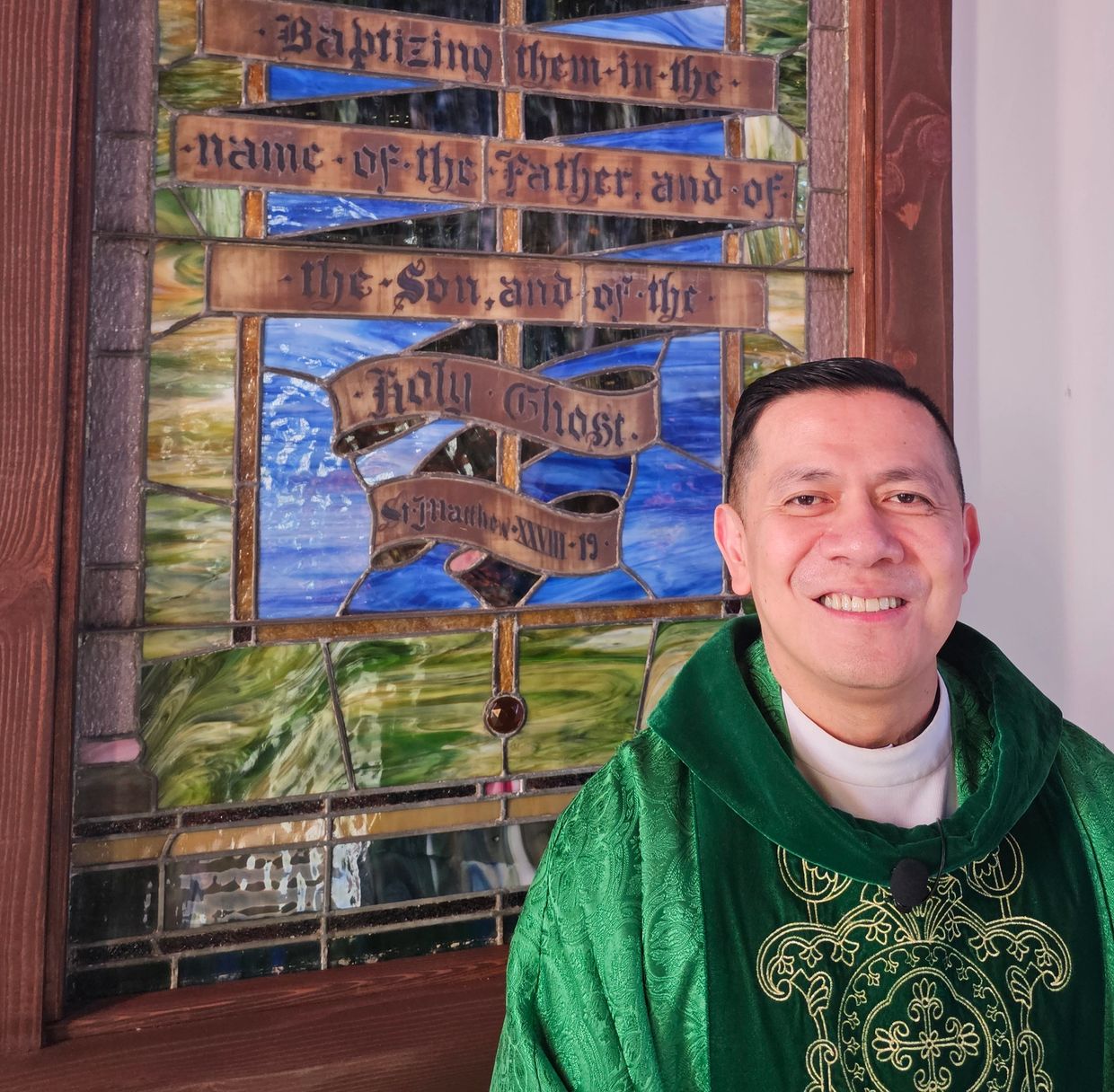 Smiling priest in green vestments next to stained glass.