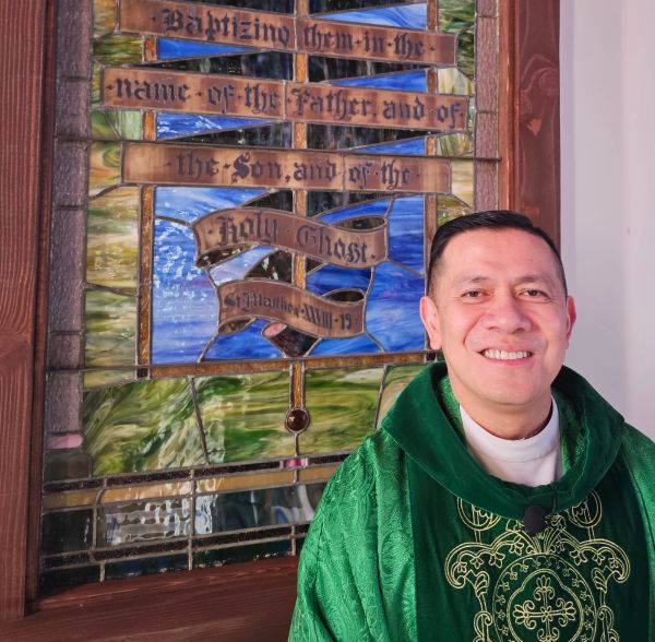 Smiling priest in green vestments next to stained glass.