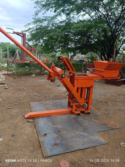 Manual orange brick-making machine on a metal sheet outdoors.